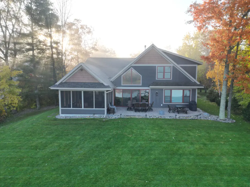 A gray-blue lake home with a screened porch and stone patio, framed by autumn trees.